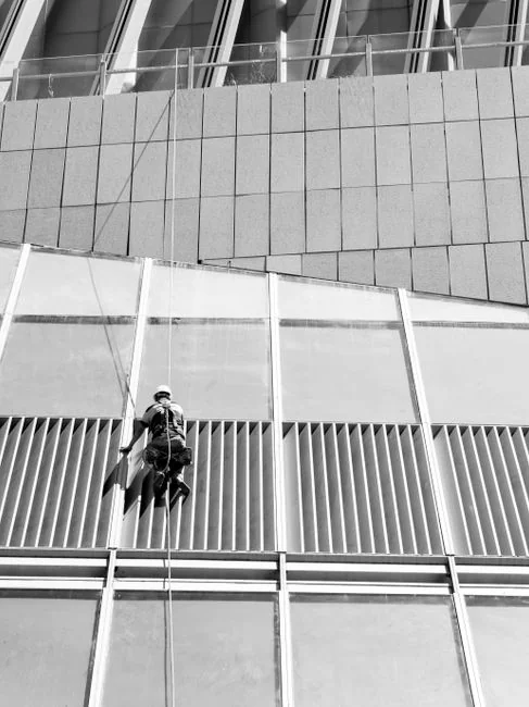 about-02 Trois laveurs de vitres suspendus à des cordes nettoient la façade d’un immeuble moderne en verre pendant la journée.